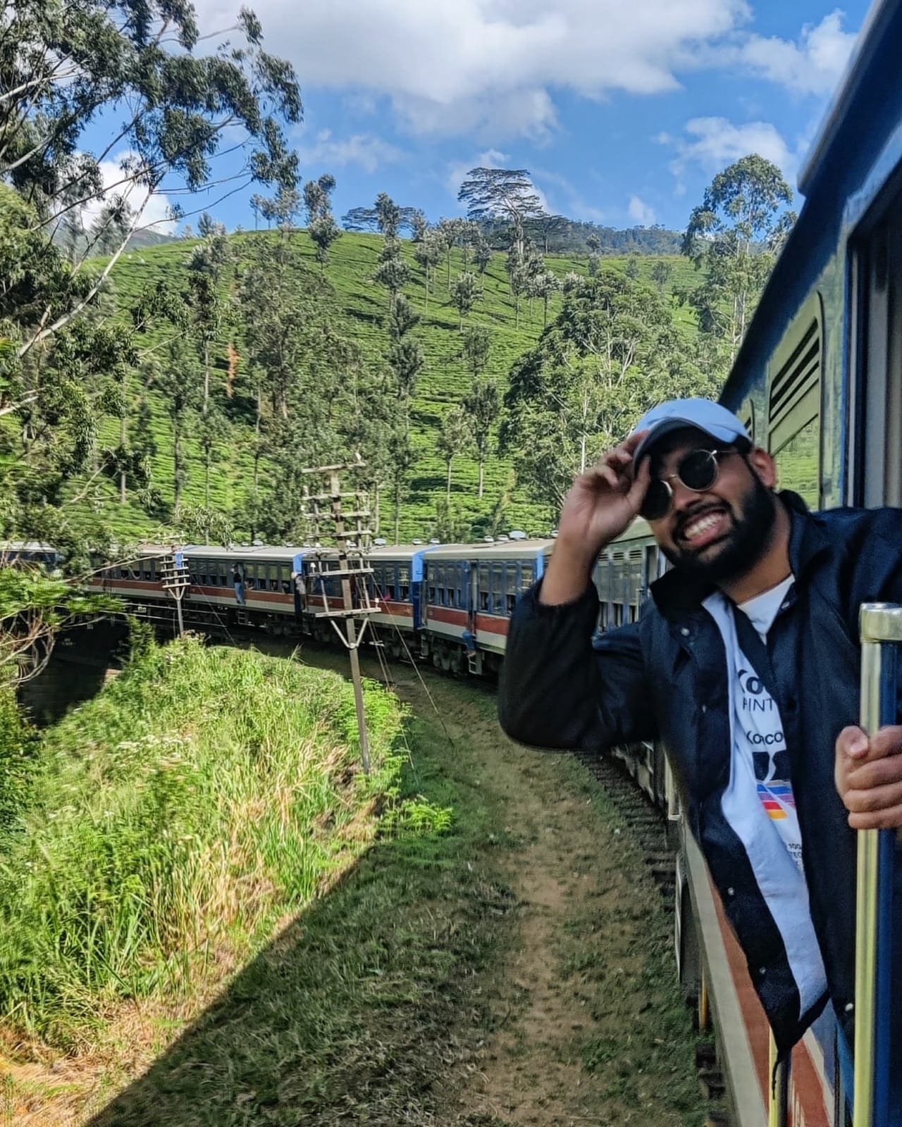 Tipping a cap from the open train door with tea fields and a forested peak behind