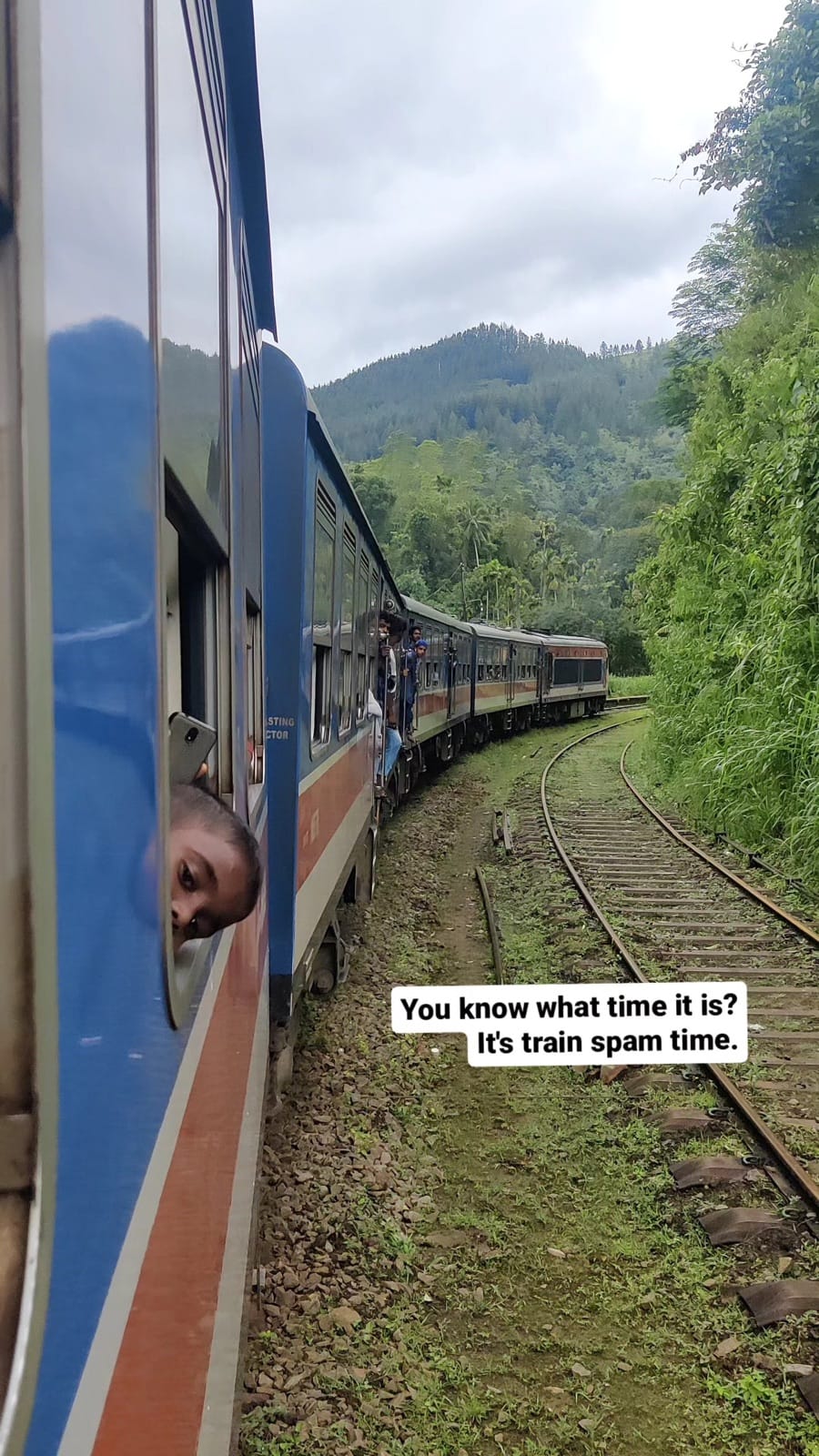 Blue Sri Lankan train curving through green hills with a kid leaning out of the window