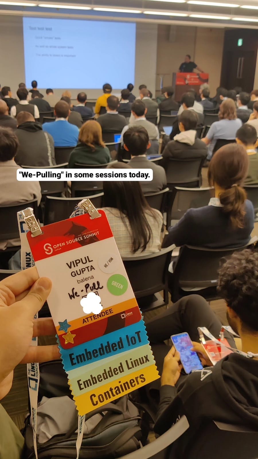 Open Source Summit Japan attendee badge with Embedded IoT, Embedded Linux and Containers ribbons held up in a packed session