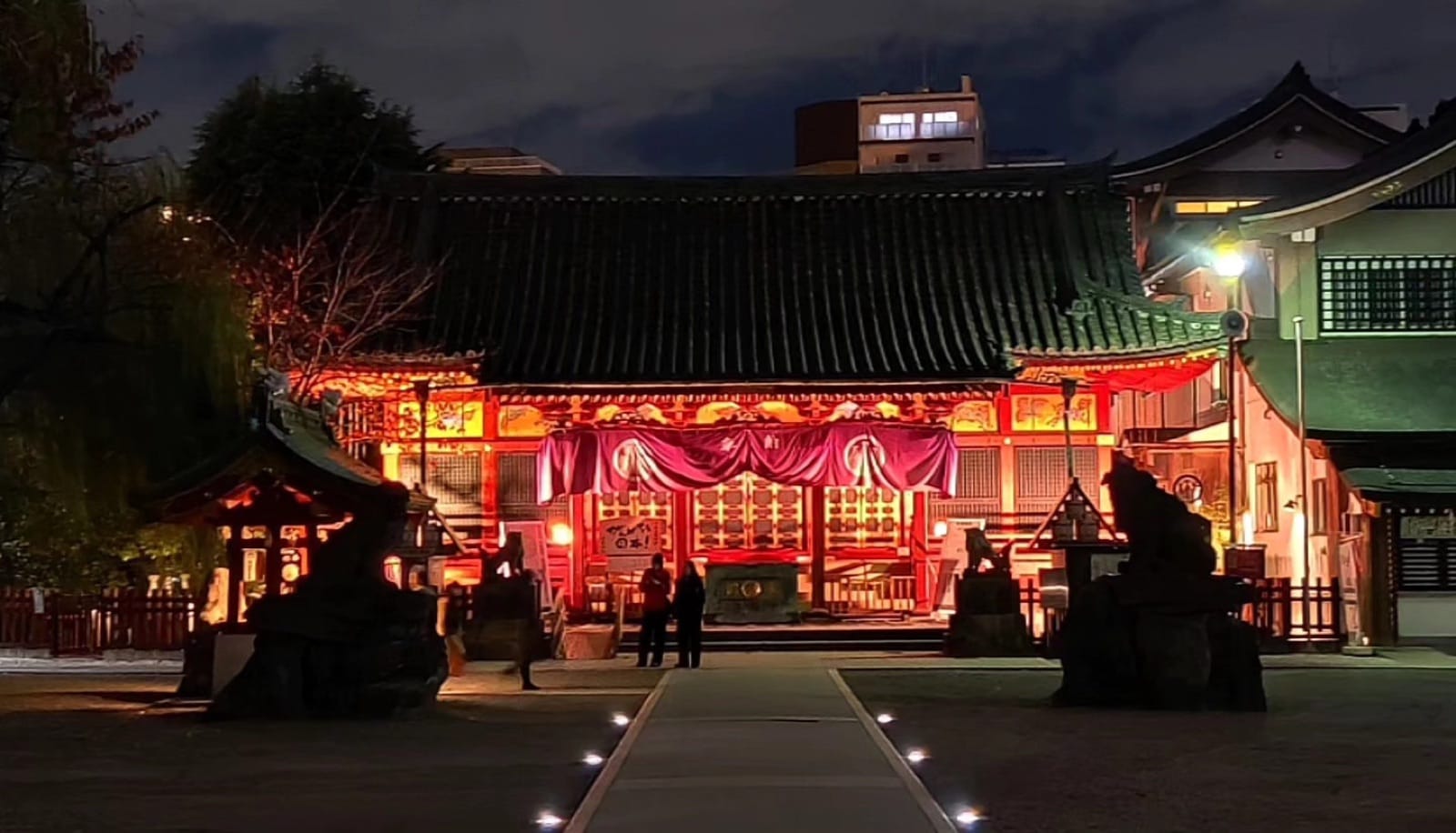 Asakusa Shrine hall glowing red behind draped curtains at dusk with stone guardians flanking the path